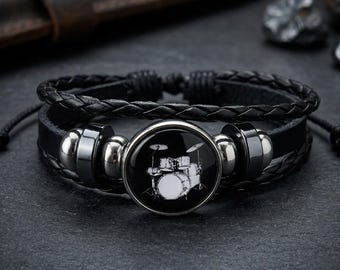 以下が含まれることがあります： 黒い革のブレスレット。編み込みバンドと、丸い銀色のセンターピースが特徴です。 センターピースには、黒い背景にドラムセットの白いイラストが描かれています。 ブレスレットには調節可能なコードが付いています。