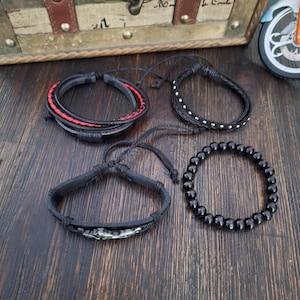 May include: Four bracelets in a variety of styles. One is black leather with red stitching, another is black with white dots, a third is black with a decorative element, and the last is made of black beads. The bracelets are on a wooden surface.