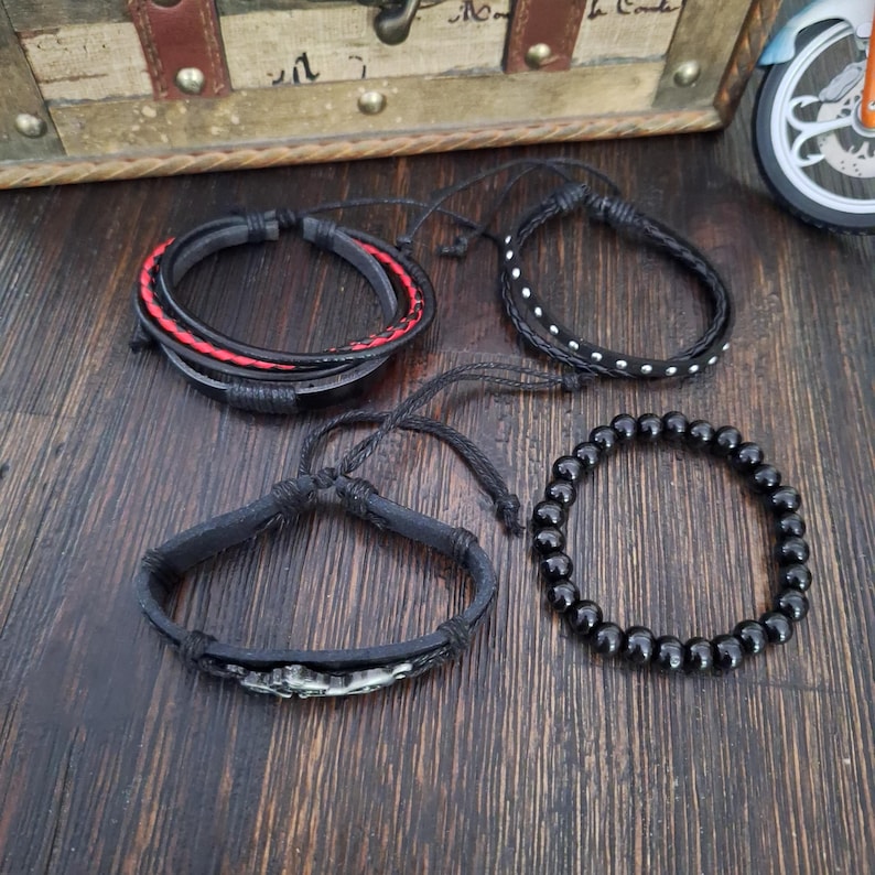 May include: Four bracelets in a variety of styles. One is black leather with red stitching, another is black with white dots, a third is black with a decorative element, and the last is made of black beads. The bracelets are on a wooden surface.