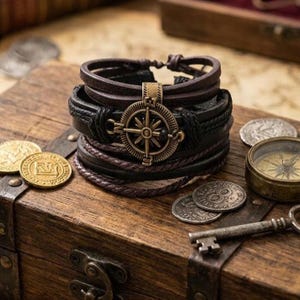 May include: A multi-layered leather bracelet with a compass charm. The bracelet features brown, black, and dark purple leather strands. The compass charm is gold-colored. The bracelet is displayed on a wooden chest with coins and a compass.