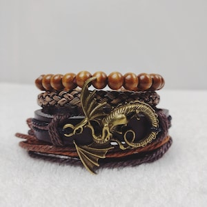 Puede incluir: Un conjunto de pulseras marrones apiladas. Incluyen cuentas de madera, cuero trenzado y correas de cuero. Un dije de drag&oacute;n de color bronce est&aacute; unido a una de las pulseras.