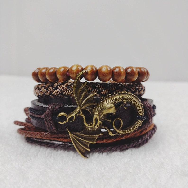 Puede incluir: Un conjunto de pulseras marrones apiladas. Incluyen cuentas de madera, cuero trenzado y correas de cuero. Un dije de drag&oacute;n de color bronce est&aacute; unido a una de las pulseras.
