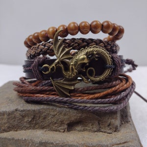 Puede incluir: Una pila de pulseras en tonos marrones y bronce. La pulsera superior tiene cuentas de madera, seguida de una pulsera trenzada, una pulsera de cuero con un dije de drag&oacute;n y una pulsera de cord&oacute;n envuelto. El dije de drag&oacute;n es bronce.