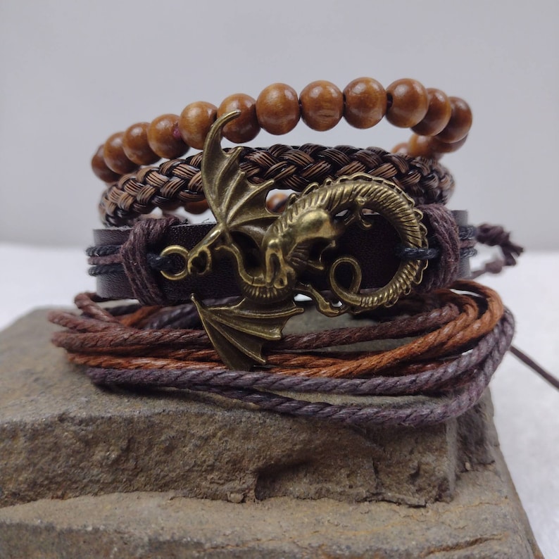 Puede incluir: Una pila de pulseras en tonos marrones y bronce. La pulsera superior tiene cuentas de madera, seguida de una pulsera trenzada, una pulsera de cuero con un dije de drag&oacute;n y una pulsera de cord&oacute;n envuelto. El dije de drag&oacute;n es bronce.