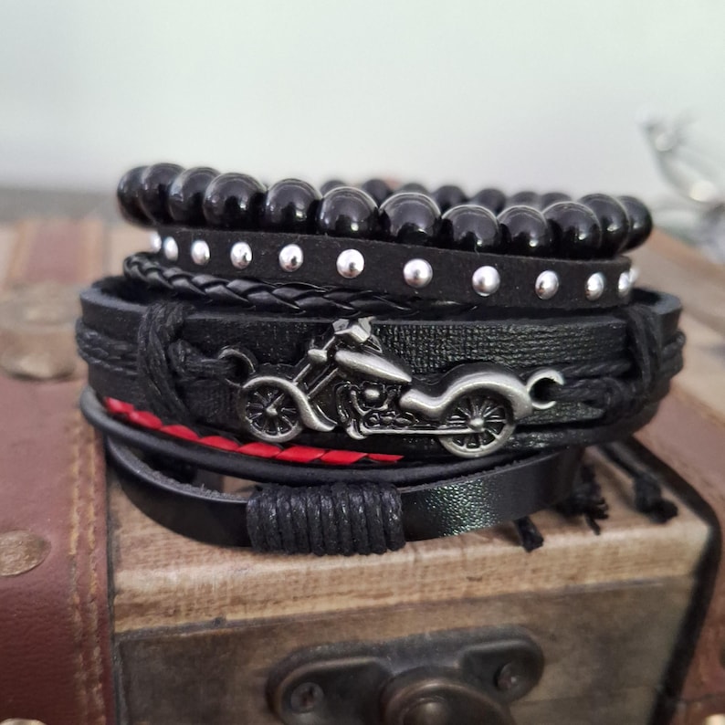 May include: A stack of black bracelets with various designs. The bracelets include beaded, studded, braided, and leather styles. A silver motorcycle charm is featured on one of the bracelets. The bracelets are stacked on a wooden surface.