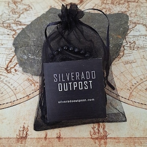 Puede incluir: Una bolsa de regalo de organza negra con cierre de cord&oacute;n, que contiene una pulsera de cuentas oscuras. Una etiqueta negra con el texto "SILVERADO OUTPOST" y la direcci&oacute;n del sitio web est&aacute; frente a la bolsa. El fondo es un mapa antiguo.