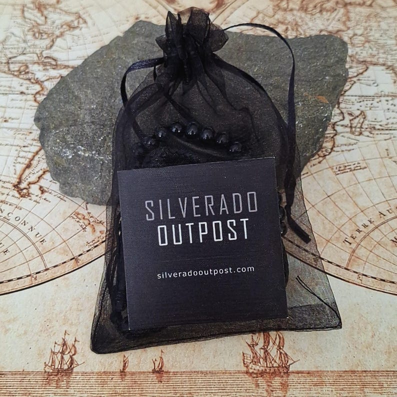 Puede incluir: Una bolsa de regalo de organza negra con cierre de cord&oacute;n, que contiene una pulsera de cuentas oscuras. Una etiqueta negra con el texto "SILVERADO OUTPOST" y la direcci&oacute;n del sitio web est&aacute; frente a la bolsa. El fondo es un mapa antiguo.