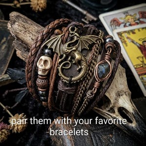 Puede incluir: Conjunto de pulseras de cuero de m&uacute;ltiples hebras con una cuenta de calavera, un dije de drag&oacute;n de bronce y una piedra envuelta en alambre. Las pulseras son en tonos marr&oacute;n y bronce, con una banda de cuero trenzado. El texto de la imagen dice: "comb&iacute;nalas con tus pulseras favoritas."