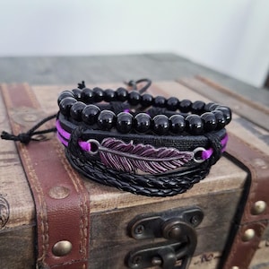 May include: A layered bracelet set featuring black beads, black leather cord, and a purple metal feather charm.
