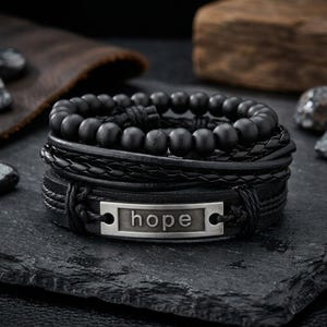 May include: A stack of black bracelets with a variety of designs. One bracelet features a series of black beads, while others have braided leather and a metal plate with the word "hope". The bracelets are displayed on a dark, textured surface.