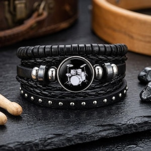 May include: Black leather bracelet with a multi-strand design. The bracelet features a round, black and white drum set graphic. Silver-toned metal accents and rivets add detail. A stylish accessory for music lovers.