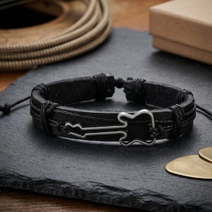 May include: Black leather bracelet with a silver guitar charm. The bracelet features a braided design and adjustable closure. A guitar pick is visible in the background. This accessory is perfect for music lovers.