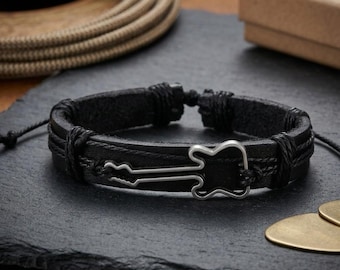 May include: Black leather bracelet with a silver guitar charm. The bracelet features a braided design and adjustable closure. A guitar pick is visible in the background. This accessory is perfect for music lovers.