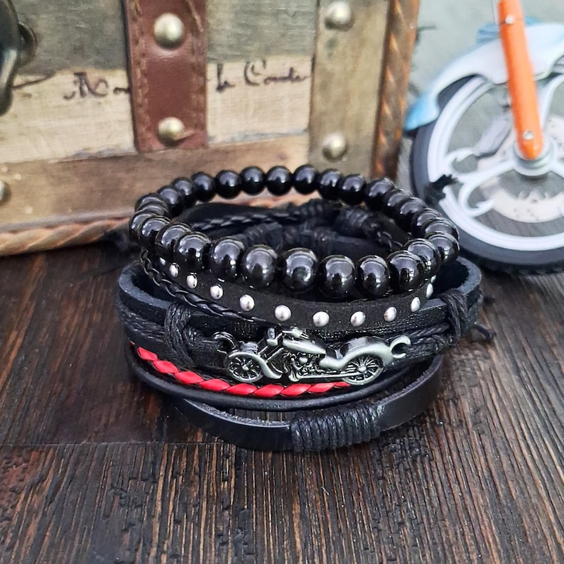 May include: A stack of black bracelets with a motorcycle charm. The bracelets include beaded, studded, and braided designs. One bracelet has a red braided accent. The bracelets are displayed on a wooden surface.