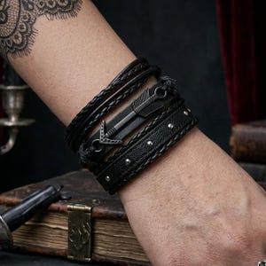 May include: A stack of black leather bracelets with metal arrow and stud accents. The bracelets are braided, studded, and feature an arrow design. The bracelets are worn on the wrist.