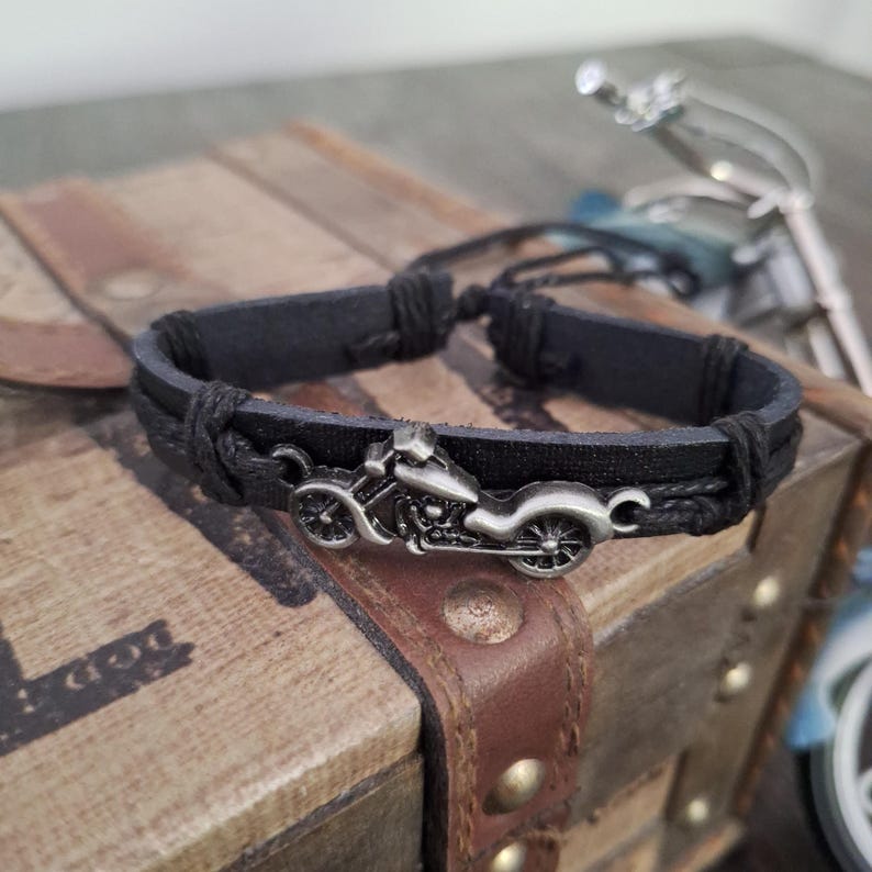 May include: Black leather bracelet featuring a silver-colored motorcycle charm. The bracelet has a braided design and adjustable closure. The motorcycle charm is detailed with wheels, a seat, and handlebars. The bracelet is displayed on a wooden surface.