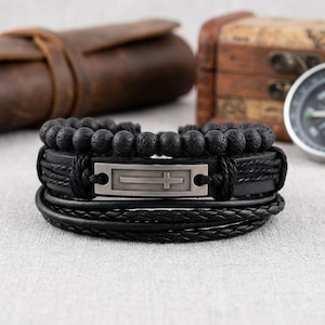 May include: A set of black bracelets. One bracelet features a series of black lava beads. Another has a silver-colored rectangular plate with a cross design. The other bracelets are made of braided and smooth black leather.