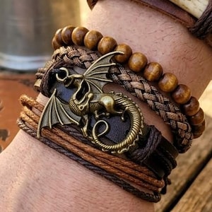 Puede incluir: Varios brazaletes marrones superpuestos, incluyendo modelos de cuero, con cuentas y trenzados. Un brazalete est&aacute; adornado con un dije de drag&oacute;n de bronce. Los brazaletes presentan diferentes tonos de marr&oacute;n.