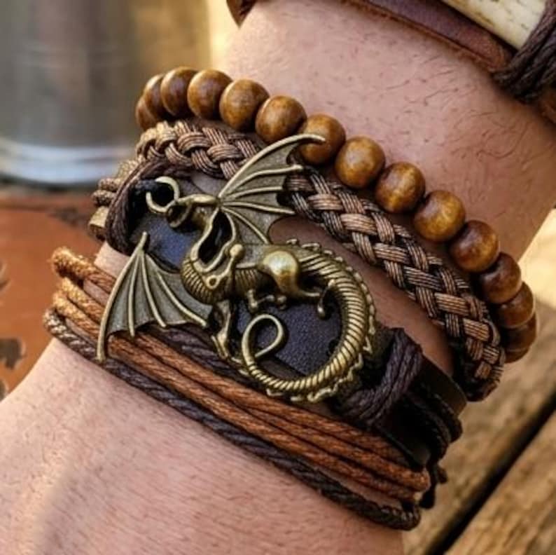 Puede incluir: Varios brazaletes marrones superpuestos, incluyendo modelos de cuero, con cuentas y trenzados. Un brazalete est&aacute; adornado con un dije de drag&oacute;n de bronce. Los brazaletes presentan diferentes tonos de marr&oacute;n.