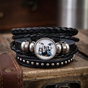 May include: Black leather bracelet with a drum set graphic. The bracelet features multiple strands of braided and studded leather, with silver-toned metal accents. The central charm displays a colorful drum kit illustration.