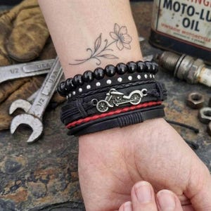May include: A stack of black leather bracelets with silver accents and a motorcycle charm. One bracelet features red stitching. The bracelets are worn on a wrist with a floral tattoo. The background includes tools and a vintage oil can.