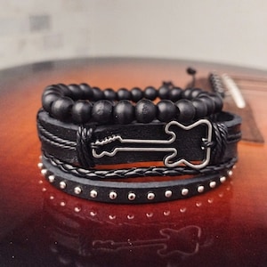 May include: A stack of black bracelets with a rock and roll theme. The bracelets include a beaded bracelet, a leather bracelet with a silver guitar outline, a braided leather bracelet, and a studded leather bracelet. The bracelets are displayed on a guitar.