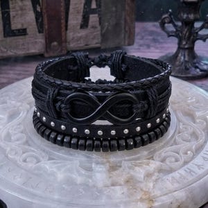 May include: A stack of black leather bracelets. The top bracelet is braided and knotted. The second bracelet features an infinity symbol. The third bracelet has silver studs. The bottom bracelet is made of black beads. The bracelets are displayed on a white, carved surface.