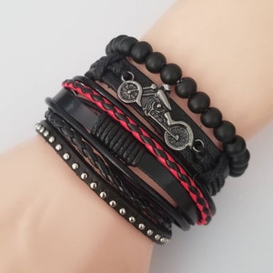 May include: A multi-layered bracelet featuring black leather straps, some with silver studs and braided details. A red braided leather strap adds a pop of color. The bracelet includes a motorcycle charm and black beaded accents.