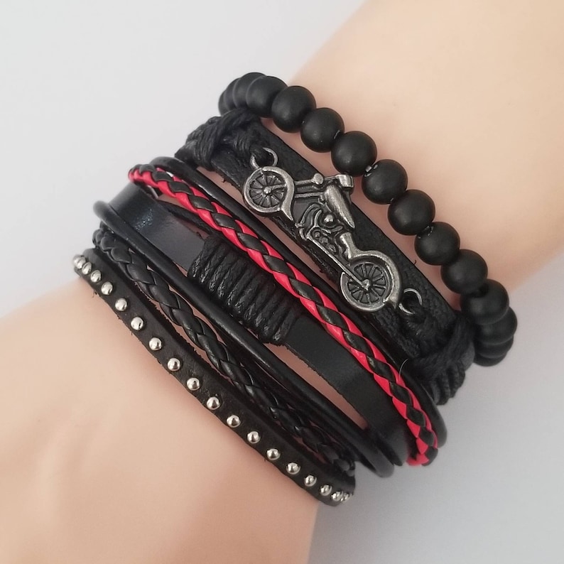 May include: A multi-layered bracelet featuring black leather straps, some with silver studs and braided details. A red braided leather strap adds a pop of color. The bracelet includes a motorcycle charm and black beaded accents.