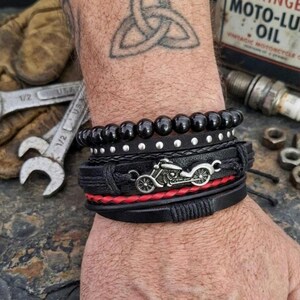 May include: A stack of black bracelets with a motorcycle charm, red accents, and studded details. The bracelets are worn on a wrist with a Celtic knot tattoo. The background includes tools and a vintage oil can.