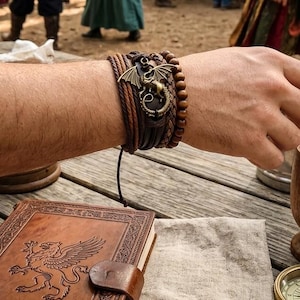 Puede incluir: Una pulsera de cuero marr&oacute;n de m&uacute;ltiples hebras con un dije de drag&oacute;n de bronce. La pulsera presenta elementos trenzados y con cuentas. Tambi&eacute;n se ve un diario encuadernado en cuero con un dise&ntilde;o de grifo.