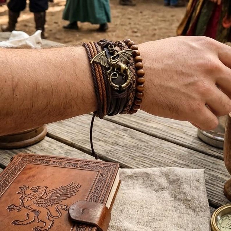 Puede incluir: Una pulsera de cuero marr&oacute;n de m&uacute;ltiples hebras con un dije de drag&oacute;n de bronce. La pulsera presenta elementos trenzados y con cuentas. Tambi&eacute;n se ve un diario encuadernado en cuero con un dise&ntilde;o de grifo.