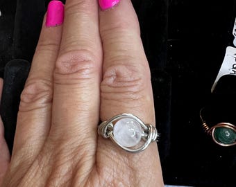 May include: A silver wire wrapped ring with a round, white gemstone. The ring is on a finger with pink nail polish.