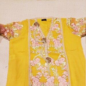 K&ouml;nnte beinhalten: Senfgelbe Tunika mit floraler Stickerei. Die Tunika hat einen V-Ausschnitt, verziert mit dekorativen Elementen und einem Mittelteil aus rosa und gr&uuml;ner Blumenstickerei. Die &Auml;rmel sind transparent mit passenden floralen Details.