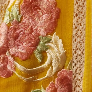 K&ouml;nnte beinhalten: Nahaufnahme eines gelben Stoffes mit gestickten rosa Blumen, wei&szlig;en Schn&ouml;rkeln und gr&uuml;nen Bl&auml;ttern. Das Design umfasst eine cremefarbene Spitzenborte an der Seite. Der Stoff hat ein strukturiertes Aussehen.