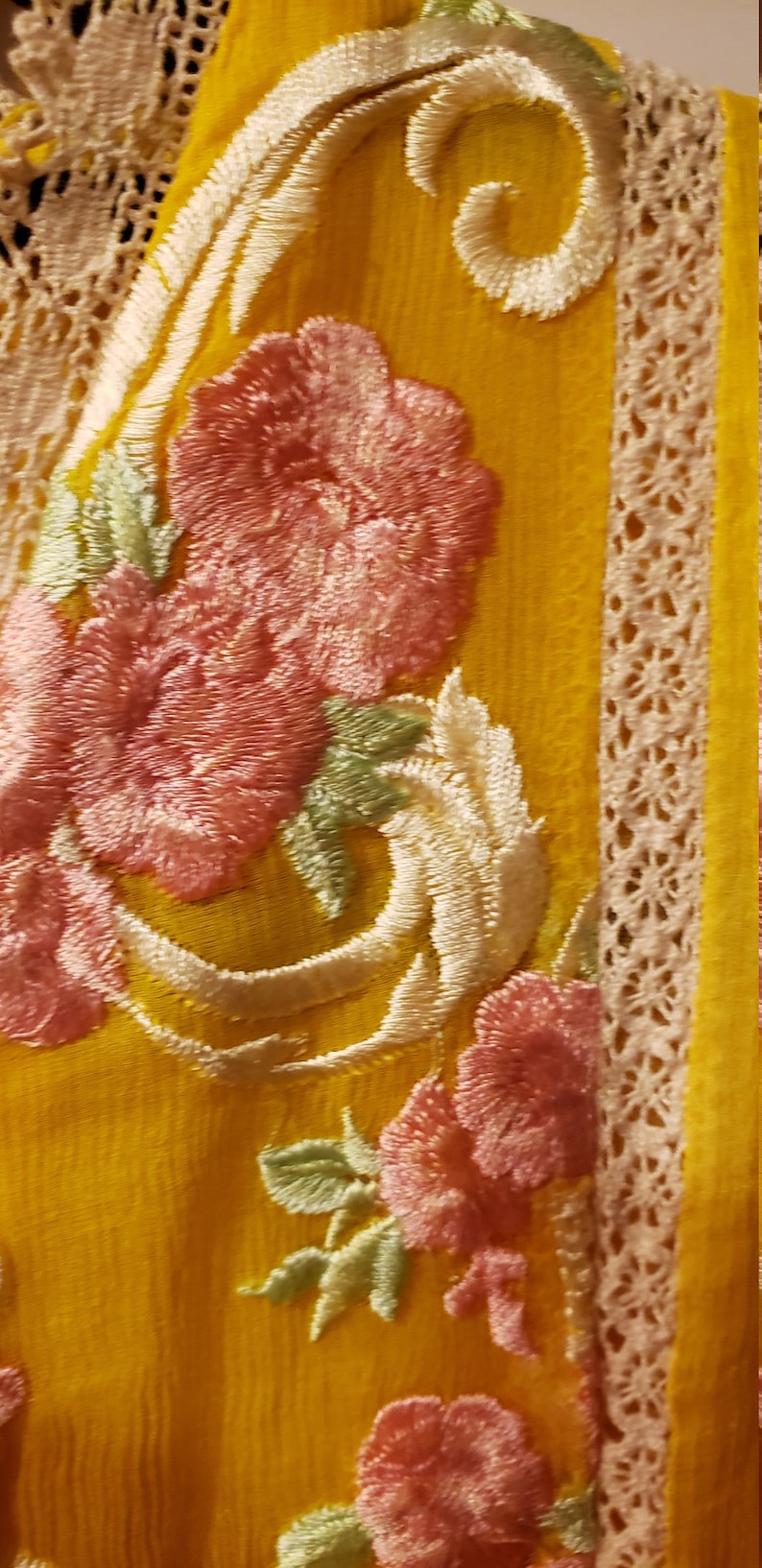 K&ouml;nnte beinhalten: Nahaufnahme eines gelben Stoffes mit gestickten rosa Blumen, wei&szlig;en Schn&ouml;rkeln und gr&uuml;nen Bl&auml;ttern. Das Design umfasst eine cremefarbene Spitzenborte an der Seite. Der Stoff hat ein strukturiertes Aussehen.