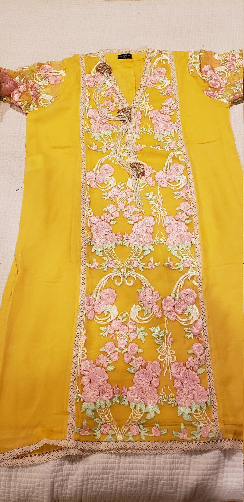 K&ouml;nnte beinhalten: Senfgelbe Tunika mit floraler Stickerei. Die Tunika hat einen V-Ausschnitt, verziert mit dekorativen Elementen und einem Mittelteil aus rosa und gr&uuml;ner Blumenstickerei. Die &Auml;rmel sind transparent mit passenden floralen Details.