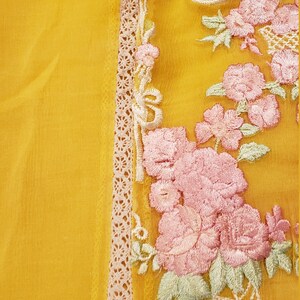 K&ouml;nnte beinhalten: Senfgelber Stoff mit aufwendiger Blumenstickerei in Rosa, Wei&szlig; und Gr&uuml;n. Das Design zeigt rosenartige Bl&uuml;ten und zarte Bl&auml;tter. Eine cremefarbene Spitzenborte umrandet den bestickten Bereich und verleiht dem Textil Eleganz.
