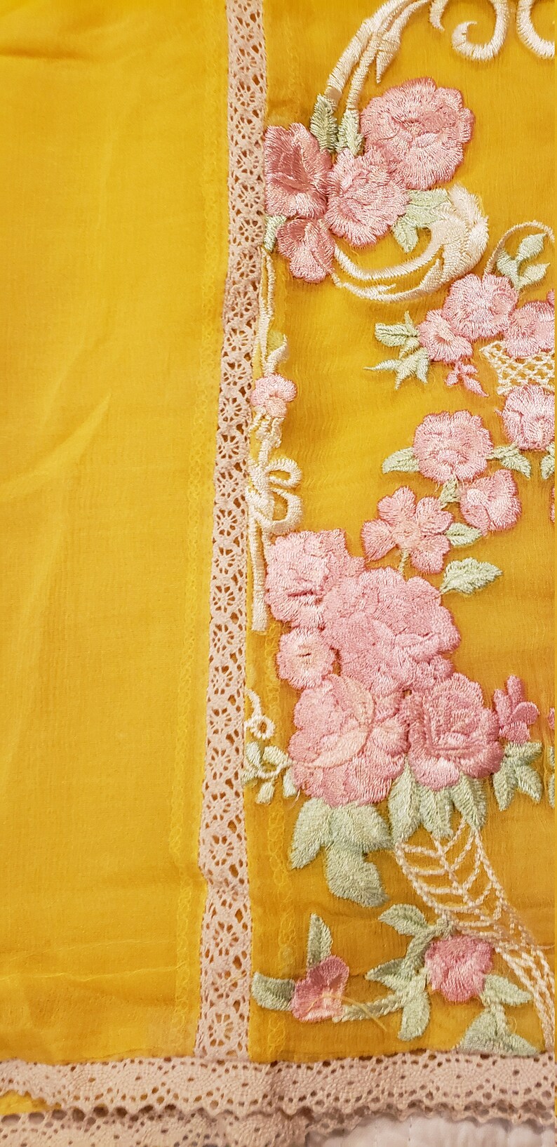 K&ouml;nnte beinhalten: Senfgelber Stoff mit aufwendiger Blumenstickerei in Rosa, Wei&szlig; und Gr&uuml;n. Das Design zeigt rosenartige Bl&uuml;ten und zarte Bl&auml;tter. Eine cremefarbene Spitzenborte umrandet den bestickten Bereich und verleiht dem Textil Eleganz.