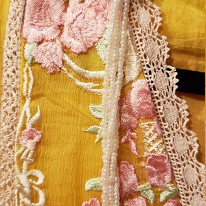 K&ouml;nnte beinhalten: Nahaufnahme eines gelben Kleidungsst&uuml;cks mit aufwendiger Blumenstickerei in Rosa und Gr&uuml;n. Das Design umfasst eine cremefarbene Spitzenborte und eine goldfarbene Verzierung mit einer Perlenkette.