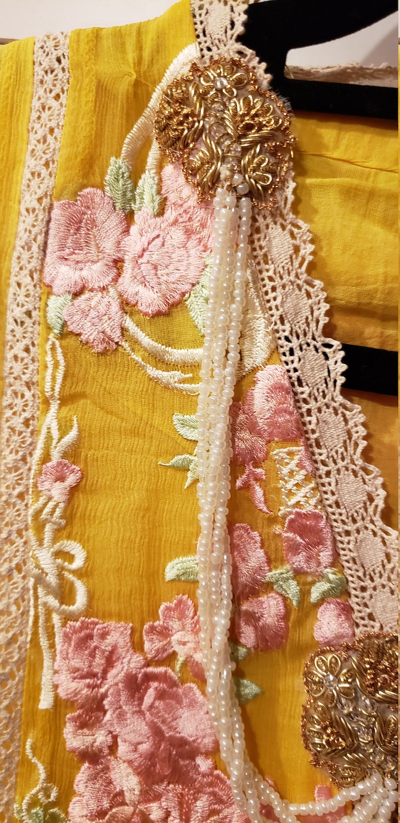 K&ouml;nnte beinhalten: Nahaufnahme eines gelben Kleidungsst&uuml;cks mit aufwendiger Blumenstickerei in Rosa und Gr&uuml;n. Das Design umfasst eine cremefarbene Spitzenborte und eine goldfarbene Verzierung mit einer Perlenkette.