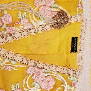 K&ouml;nnte beinhalten: Senfgelber Stoff mit rosa und wei&szlig;er Blumenstickerei. Das Kleidungsst&uuml;ck hat eine Spitzenborte und eine goldene Brosche. Das Etikett lautet "AGHA NOOR Small".