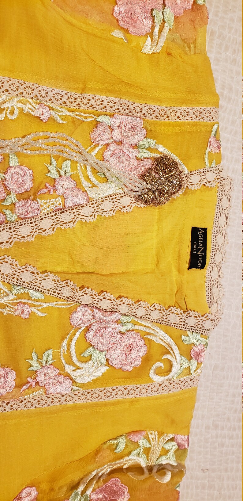 K&ouml;nnte beinhalten: Senfgelber Stoff mit rosa und wei&szlig;er Blumenstickerei. Das Kleidungsst&uuml;ck hat eine Spitzenborte und eine goldene Brosche. Das Etikett lautet "AGHA NOOR Small".