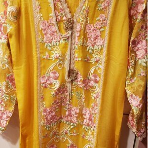 K&ouml;nnte beinhalten: Senfgelbe Tunika mit rosa und gr&uuml;ner Blumenstickerei. Das lang&auml;rmlige Oberteil hat einen V-Ausschnitt, Spitzenbesatz und dekorative Verzierungen. Die Marke "Aisha Noor" ist sichtbar.