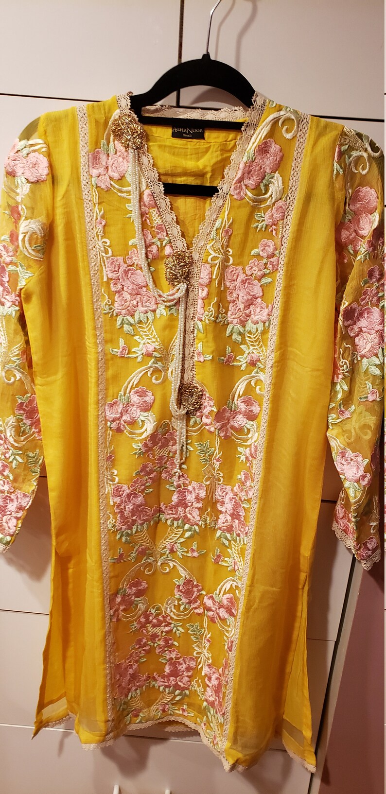 K&ouml;nnte beinhalten: Senfgelbe Tunika mit rosa und gr&uuml;ner Blumenstickerei. Das lang&auml;rmlige Oberteil hat einen V-Ausschnitt, Spitzenbesatz und dekorative Verzierungen. Die Marke "Aisha Noor" ist sichtbar.