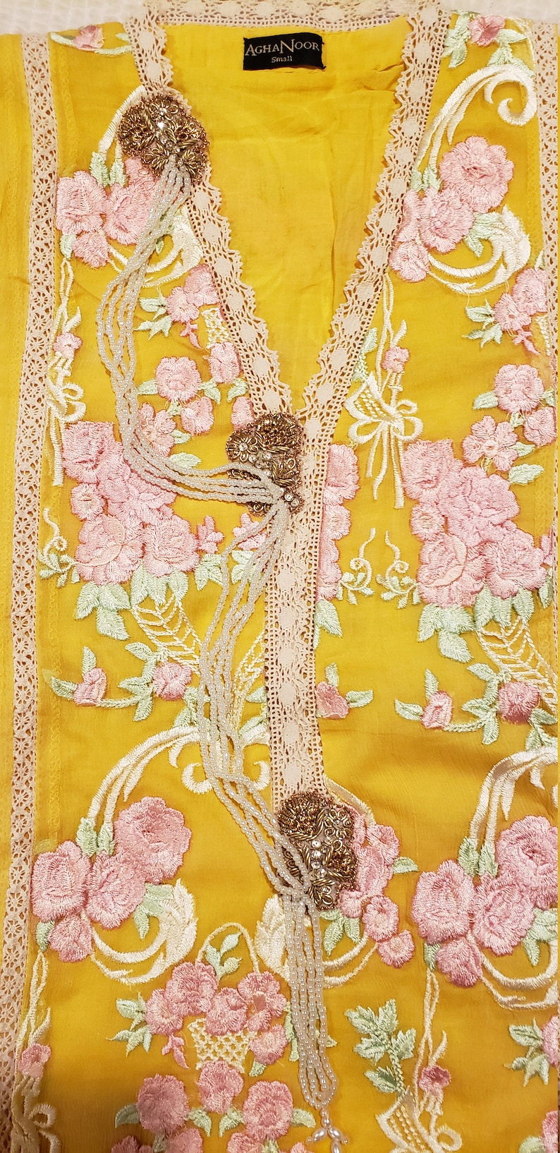 K&ouml;nnte beinhalten: Senfgelbe Tunika mit rosa Blumenstickerei und cremefarbenem Spitzenbesatz. Die Tunika hat einen V-Ausschnitt mit dekorativem Perlenbesatz. Der Markenname "AGHANOOR" steht auf dem Etikett.