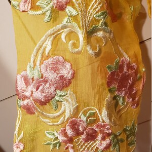 K&ouml;nnte beinhalten: Nahaufnahme eines transparenten, gelben Stoffes mit aufwendiger Blumenstickerei. Die Stickerei zeigt rosa und gr&uuml;ne Blumen mit wei&szlig;en Details. Der Stoff ist drapiert und zeigt die zarte Textur und das Design.