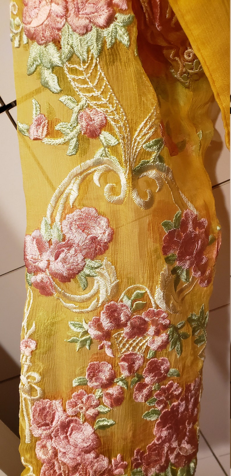 K&ouml;nnte beinhalten: Nahaufnahme eines transparenten, gelben Stoffes mit aufwendiger Blumenstickerei. Die Stickerei zeigt rosa und gr&uuml;ne Blumen mit wei&szlig;en Details. Der Stoff ist drapiert und zeigt die zarte Textur und das Design.