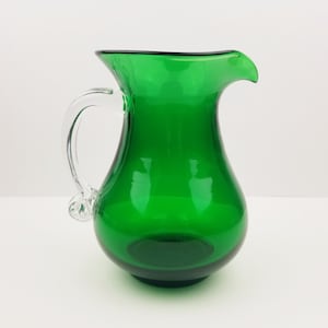 May include: A green glass pitcher with a clear glass handle. The pitcher has a rounded body and a wide mouth.
