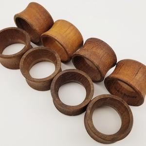 May include: A collection of eight wooden napkin rings. The rings are a warm brown color, with a smooth, polished finish. The design features a flared, curved shape. The napkin rings are arranged on a white surface.